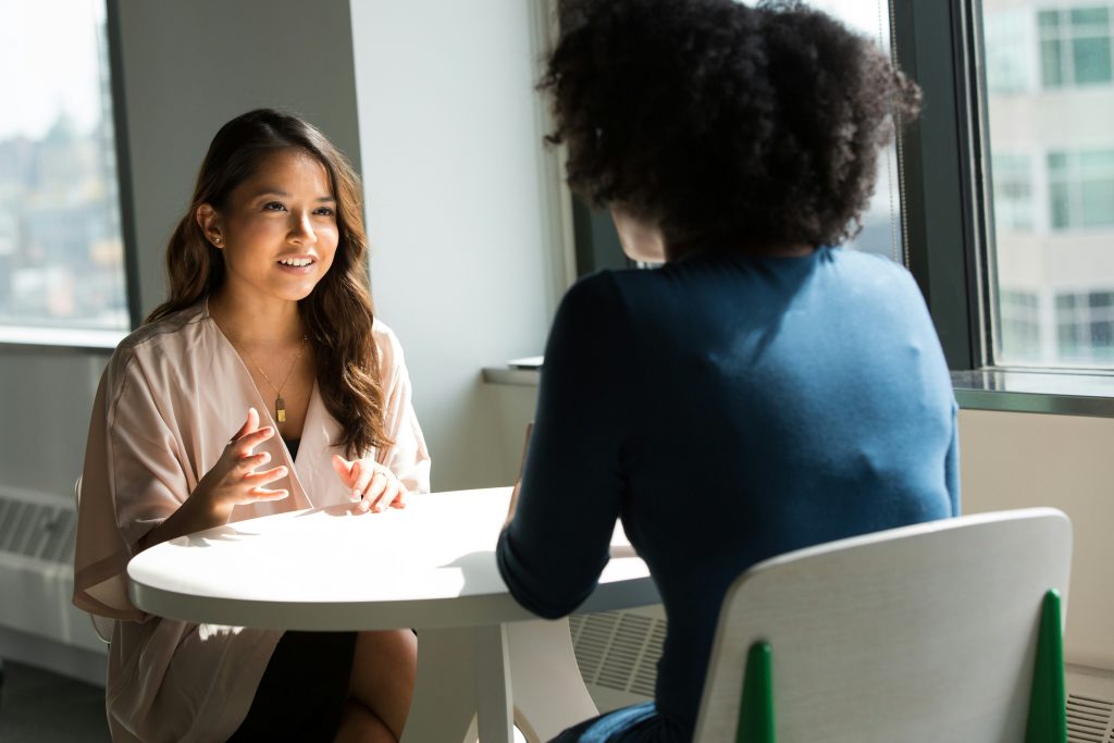2 women sitting down at a small table chatting. You can see buildings out the window behind them.