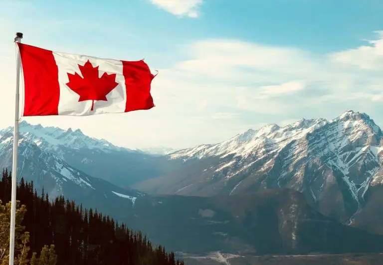 A landscape picture of snowy mountains at a distance, pine trees and a flag pole with the Canadian Flag flying in midair.